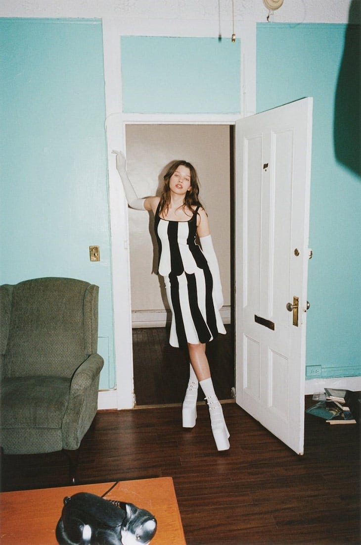 Indoor fashion shot of a woman in a black-and-white striped dress and white boots leaning in a doorway with a vintage phone on a table.