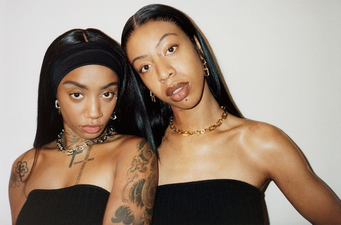 Two dark-skinned women pose indoors wearing black tops, chain necklaces, and subtle makeup, against a neutral background in a medium shot.