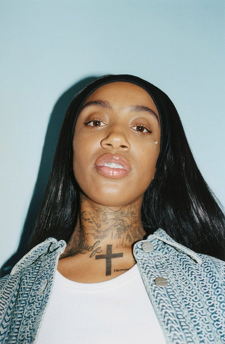 Close-up of a young adult Black woman with long black hair, wearing a white top and jean jacket, against a light blue background.