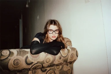 Ever Anderson wearing black eyeglasses and long black opera gloves while leaning over a patterned armchair against a white wall.