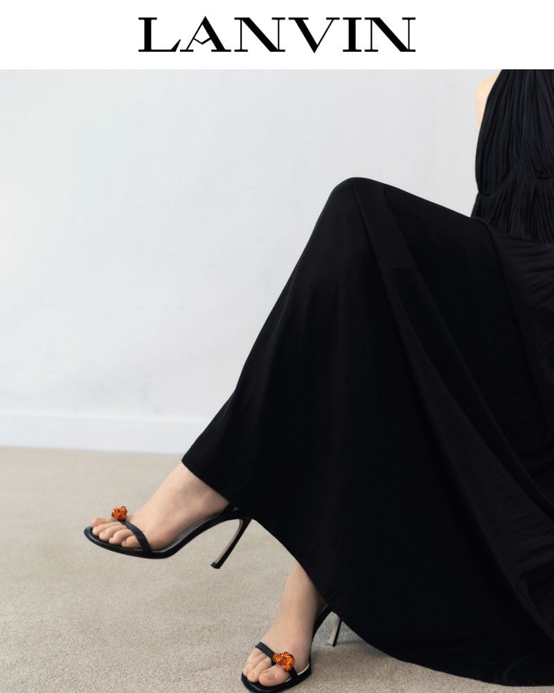 Black high-heeled sandals with floral embellishments on a woman's feet, complemented by a flowing black dress, and the 'LANVIN' logo at the top.