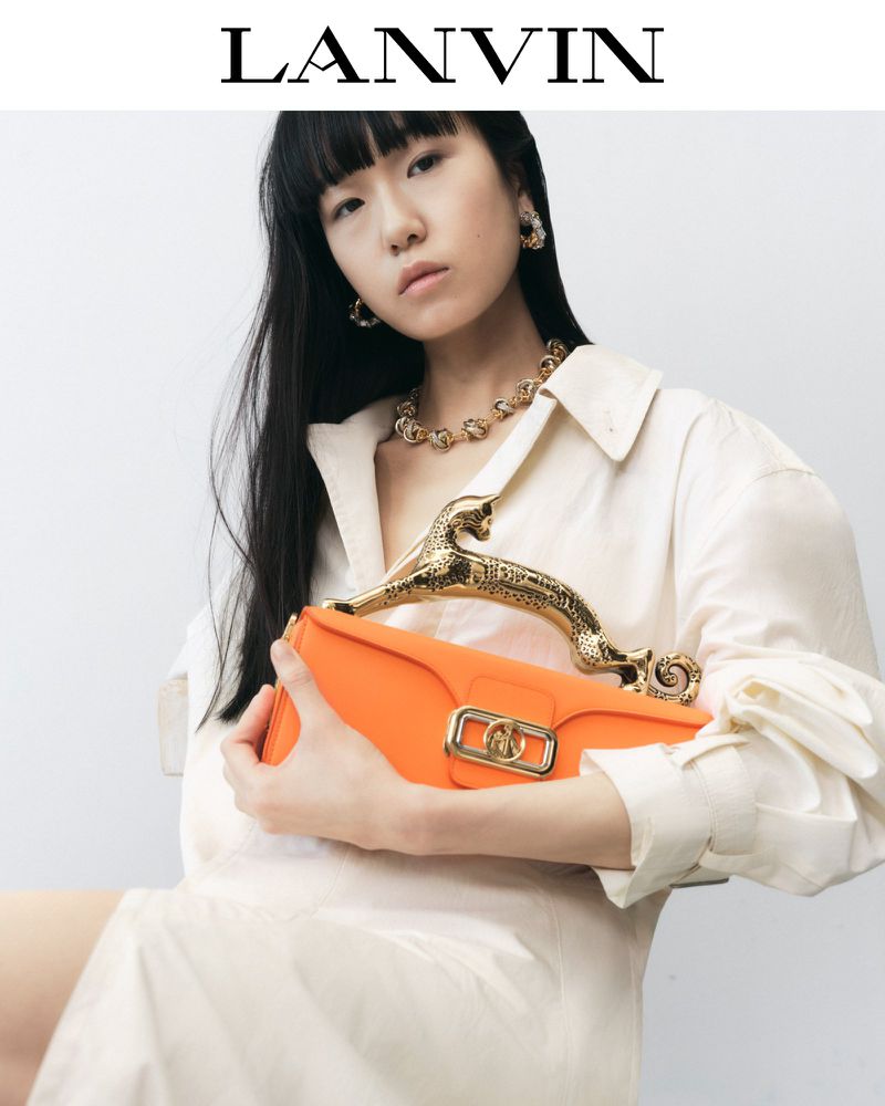 A person with dark hair poses with an orange handbag with a metallic handle and lock detail, against a plain off-white background.