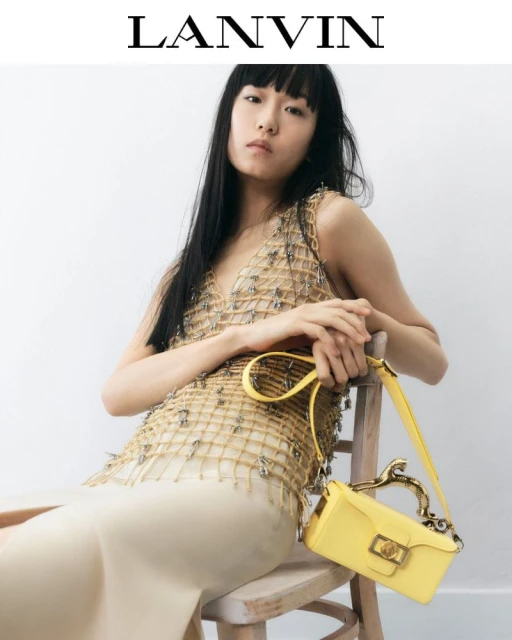 An Asian woman models for Lanvin, seated on a wooden chair with a yellow handbag against a pale white background. Fashion editorial.