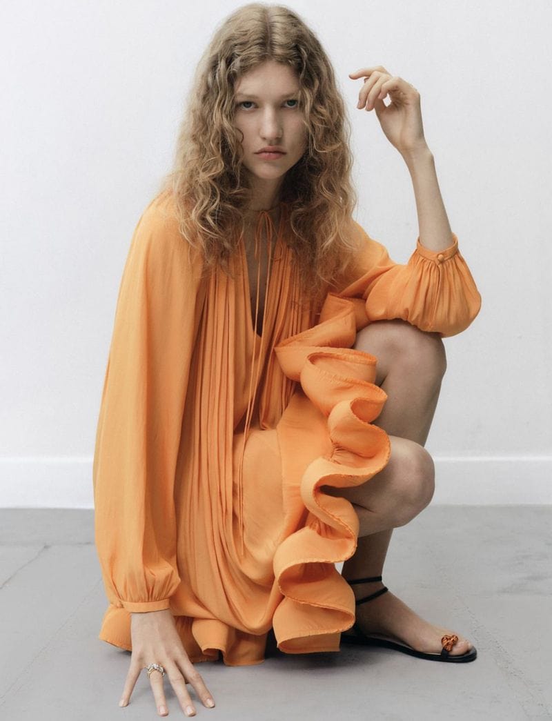 Woman in orange ruffle dress crouching in a studio. Indoor fashion shot with soft light. Curly hair and ankle-strap sandals.