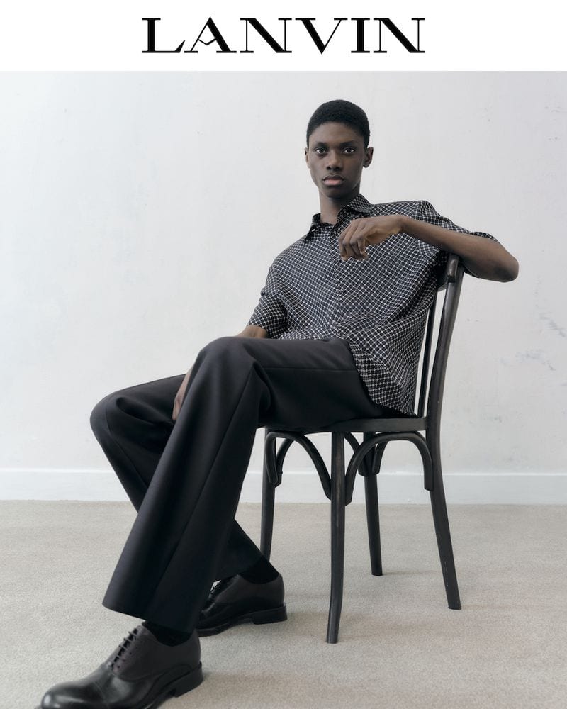 A Black male model sits in a black chair wearing a polka dot button-down shirt, wide-legged black pants, and black dress shoes for LANVIN.