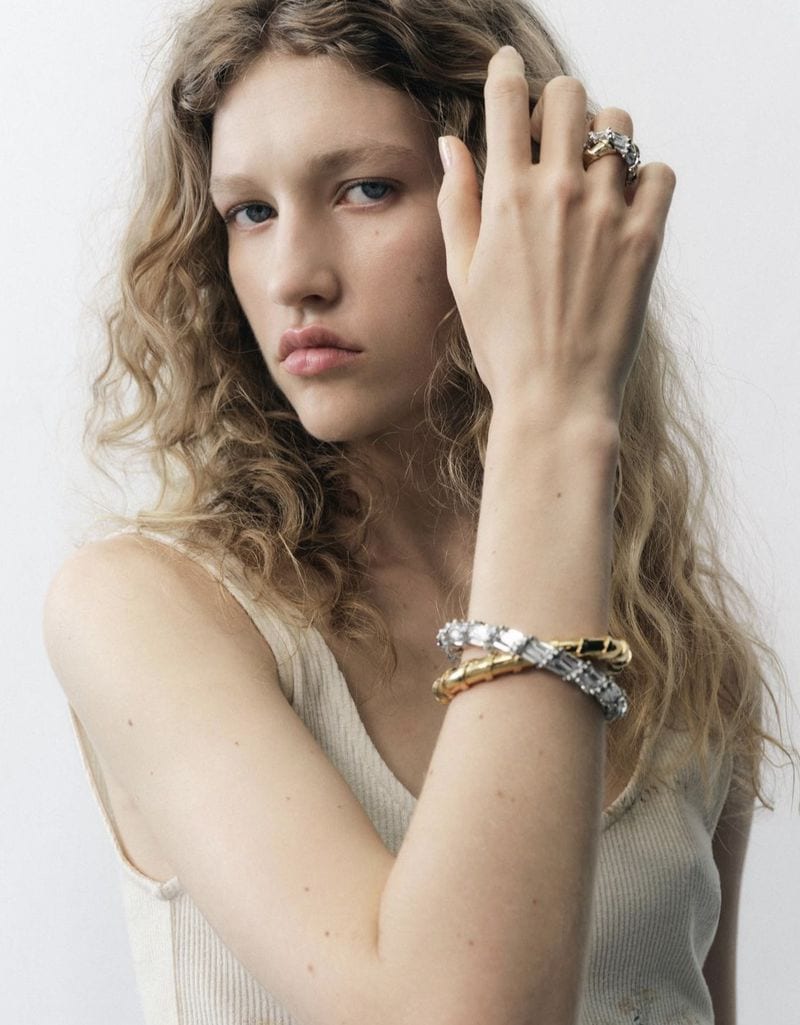 Close-up of a fair-skinned young adult female model wearing gold and crystal bracelets and rings against a light neutral backdrop.