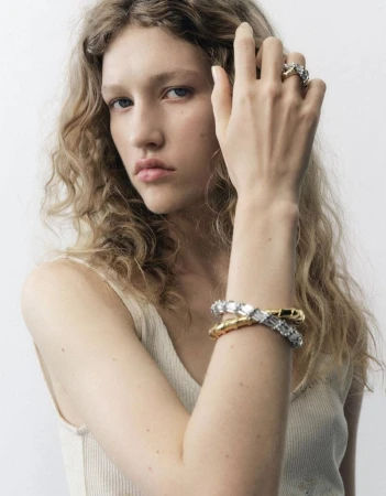 Close-up of a fair-skinned young adult female model wearing gold and crystal bracelets and rings against a light neutral backdrop.