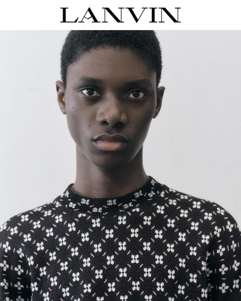 Close-up portrait of a black female model wearing a patterned black sweater, shot in a photo studio with a white backdrop and logo text.
