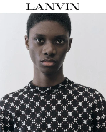 Close-up portrait of a black female model wearing a patterned black sweater, shot in a photo studio with a white backdrop and logo text.