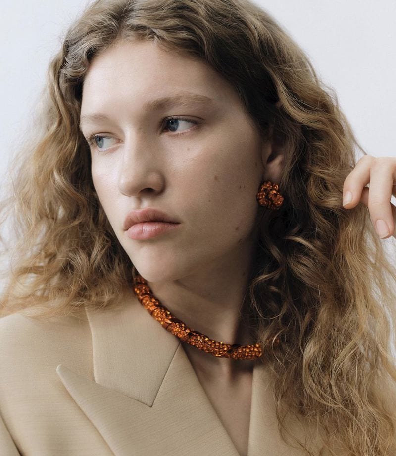 Portrait of an adult woman with wavy blonde hair, wearing a beige blazer and an orange beaded necklace and matching earrings against a white background.