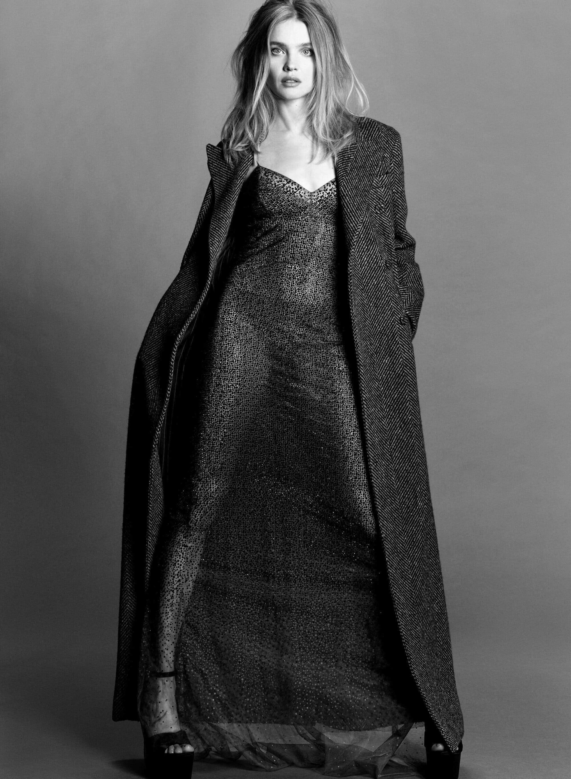 Black-and-white portrait of a blonde woman in a full-length, sequined dress and long coat, standing in a photo studio against a grey backdrop.