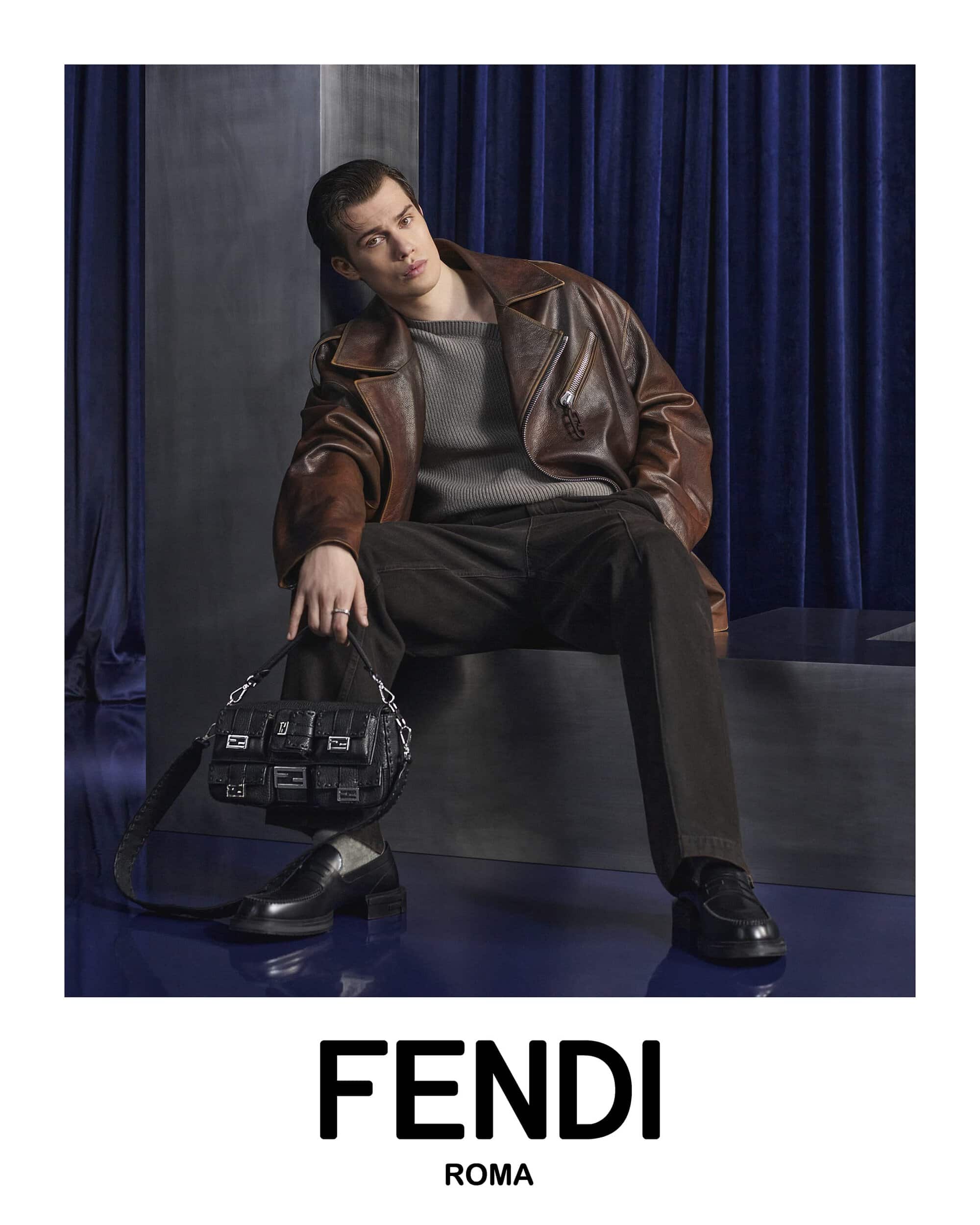 A model poses indoors wearing a brown leather jacket, grey sweater, dark pants, black shoes, and a black leather Fendi handbag.