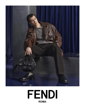 A model poses indoors wearing a brown leather jacket, grey sweater, dark pants, black shoes, and a black leather Fendi handbag.