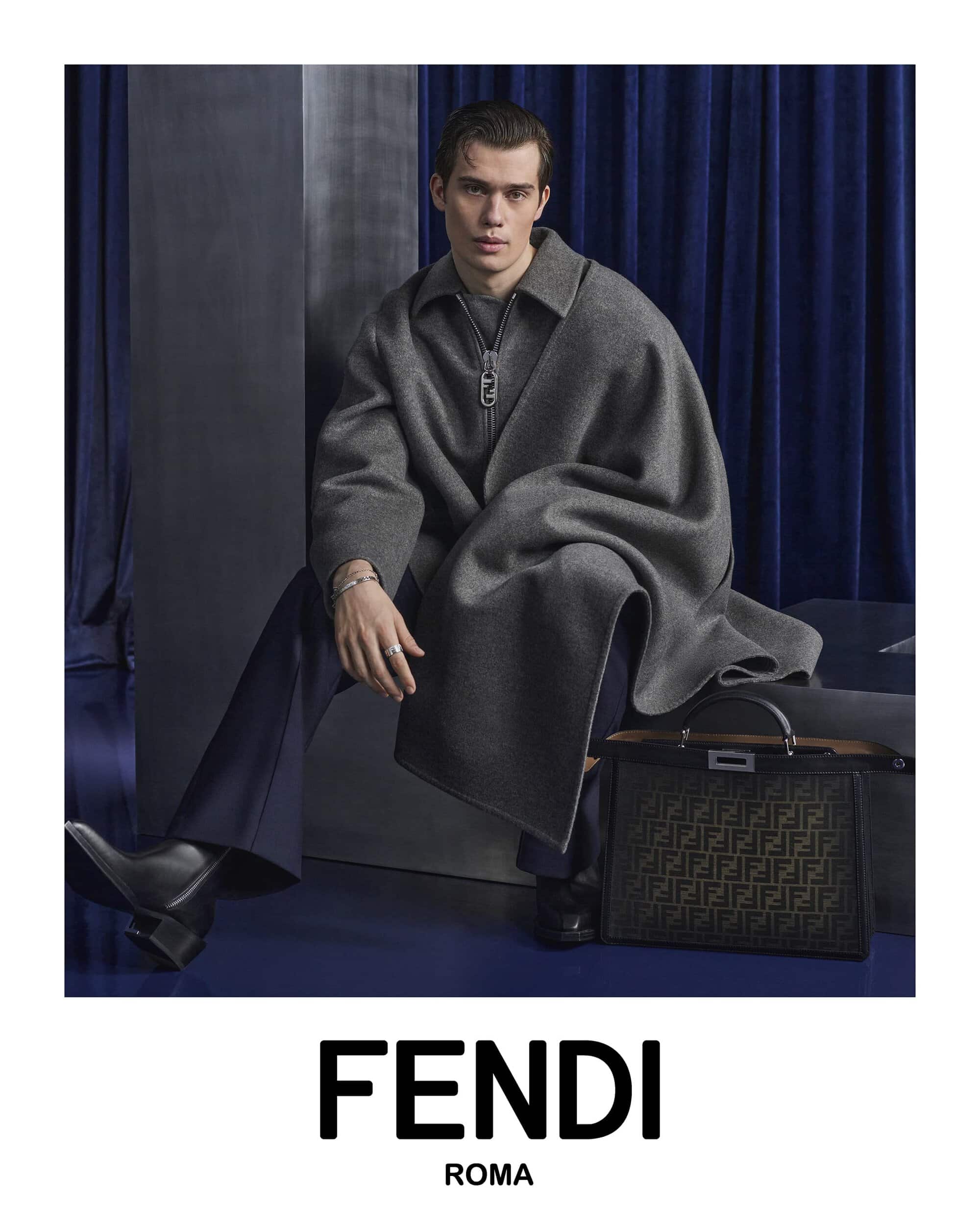 Man in a gray overcoat poses indoors next to a patterned handbag with a blue curtain background for a fashion advertisement.