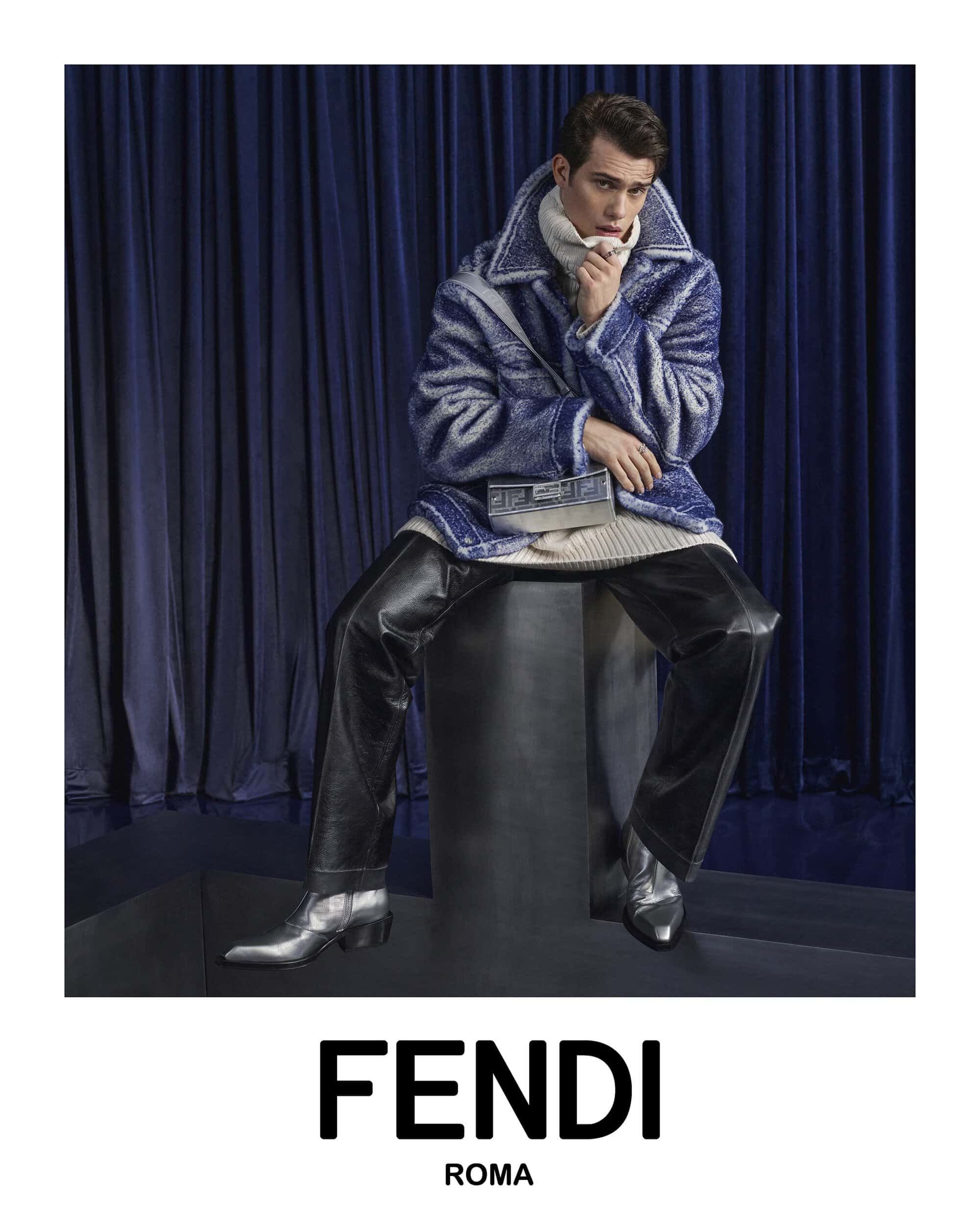 Man in blue and white coat seated in front of dark blue drapes. Wearing black pants and silver boots. Holding silver clutch.