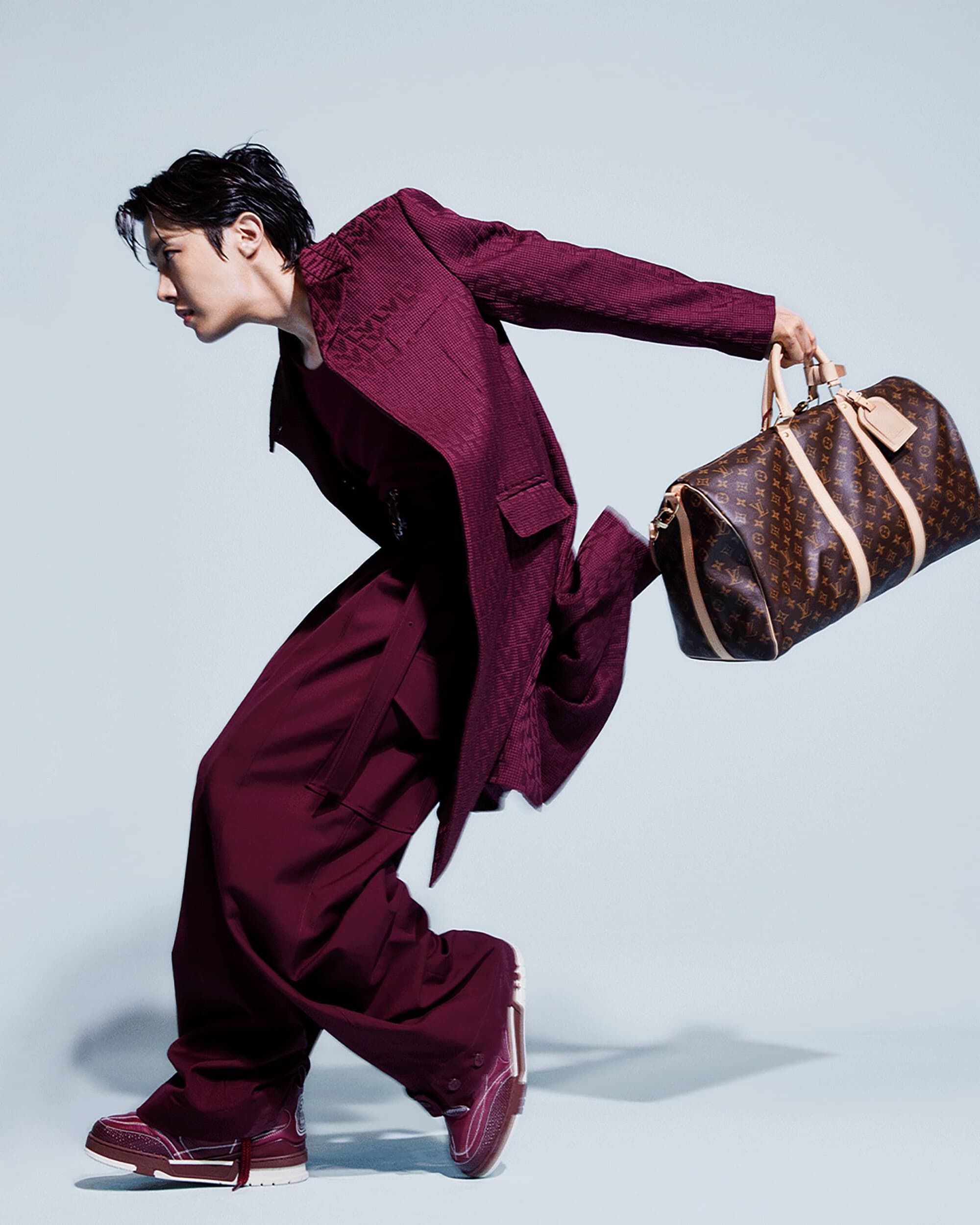 An Asian male model in a maroon suit holds a Louis Vuitton duffel bag against a light blue background in a studio setting.