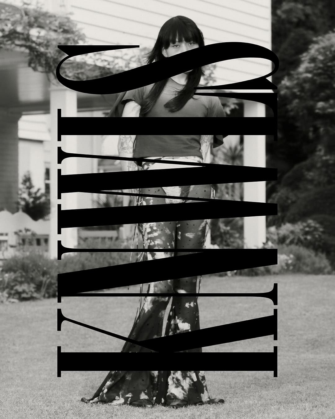 Monochrome outdoor portrait of a person in a t-shirt and tie-dye pants standing in front of a house surrounded by a green lawn.