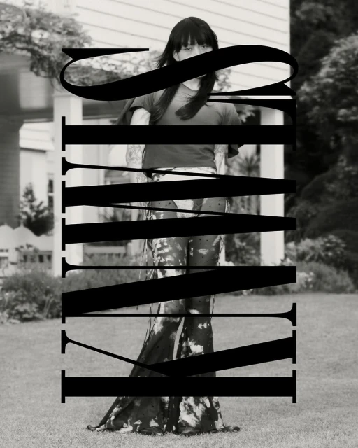Monochrome outdoor portrait of a person in a t-shirt and tie-dye pants standing in front of a house surrounded by a green lawn.
