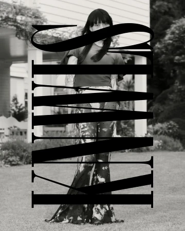 Monochrome outdoor portrait of a person in a t-shirt and tie-dye pants standing in front of a house surrounded by a green lawn.