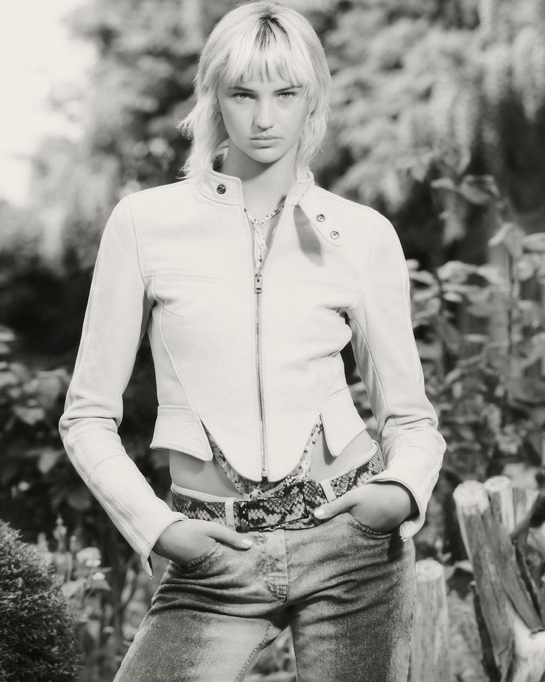 Model wearing a cropped white leather jacket and patterned belt outdoors in a black and white portrait.