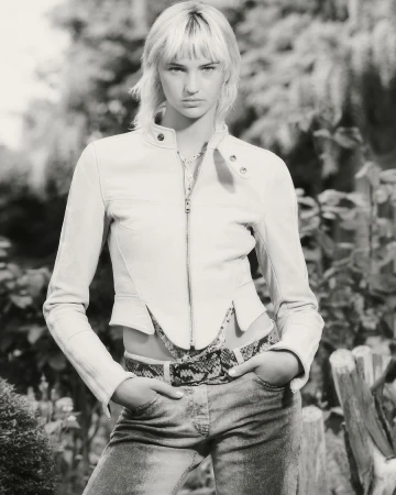 Model wearing a cropped white leather jacket and patterned belt outdoors in a black and white portrait.