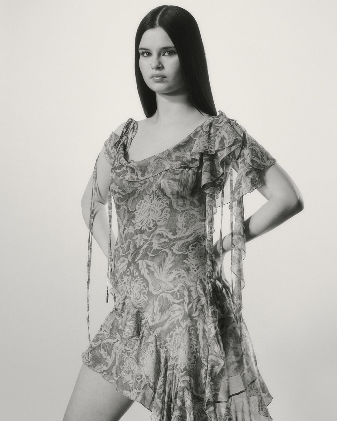 Model posing with hands on hips wearing a patterned ruffle dress against a plain studio background in monochrome.