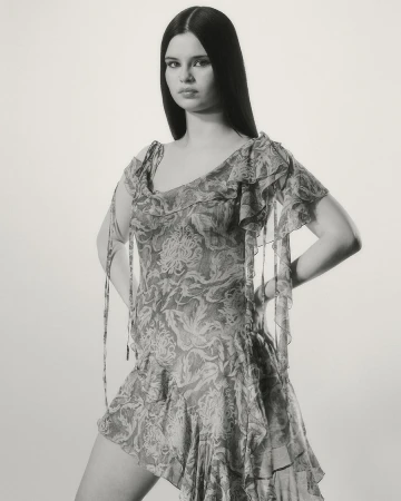 Model posing with hands on hips wearing a patterned ruffle dress against a plain studio background in monochrome.