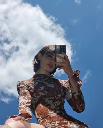 Eye-level upward shot of a woman in a patterned dress taking a selfie against a bright blue sky with scattered white clouds.