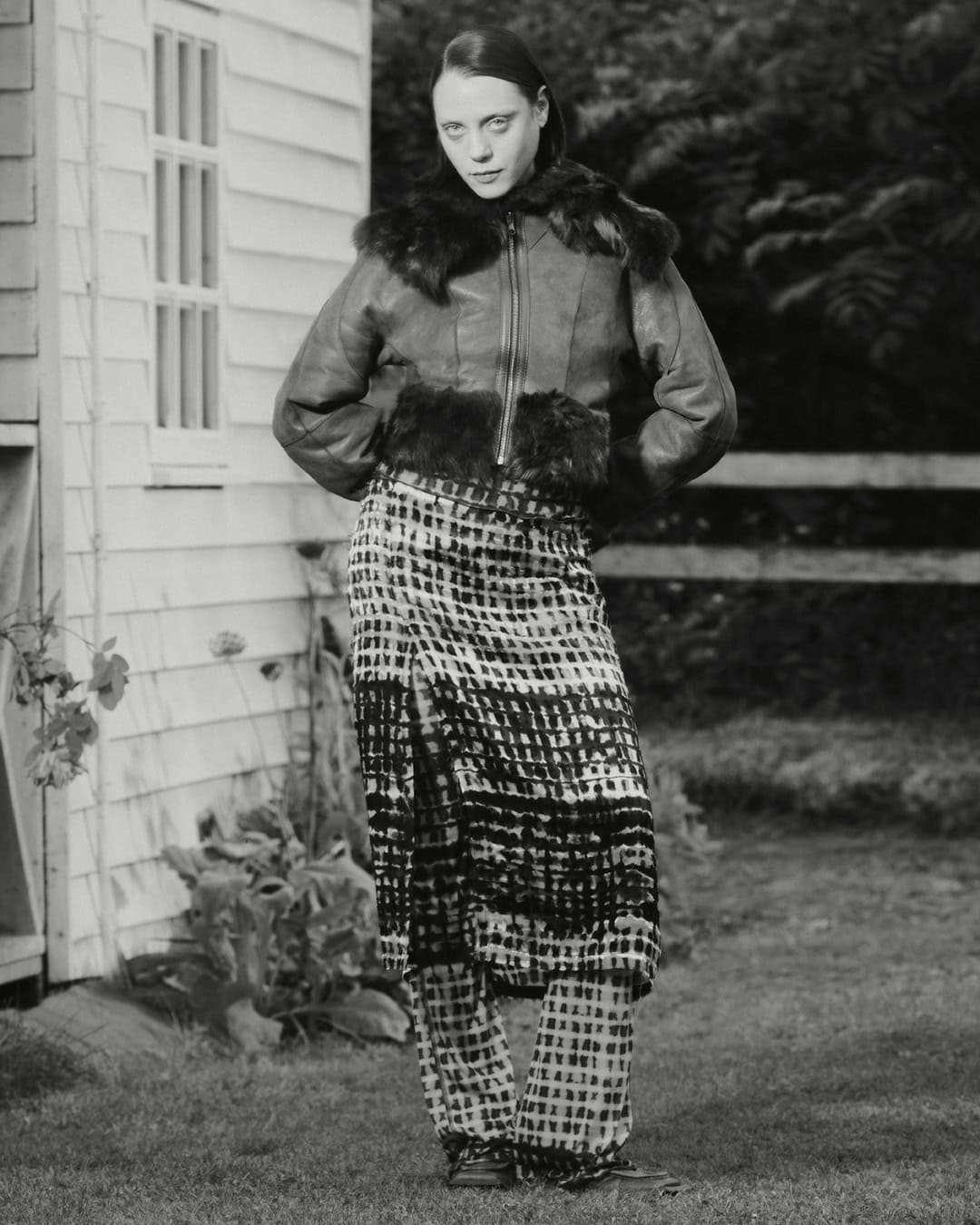 A fashion portrait of an adult female in a fur-trimmed jacket and patterned skirt and pants posing in front of a weathered building.