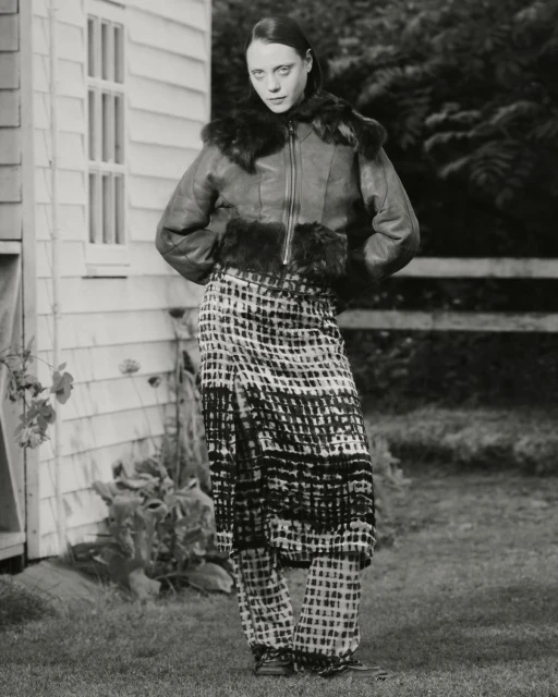 A fashion portrait of an adult female in a fur-trimmed jacket and patterned skirt and pants posing in front of a weathered building.