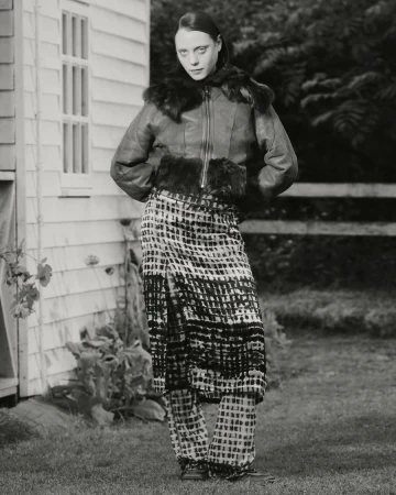 A fashion portrait of an adult female in a fur-trimmed jacket and patterned skirt and pants posing in front of a weathered building.