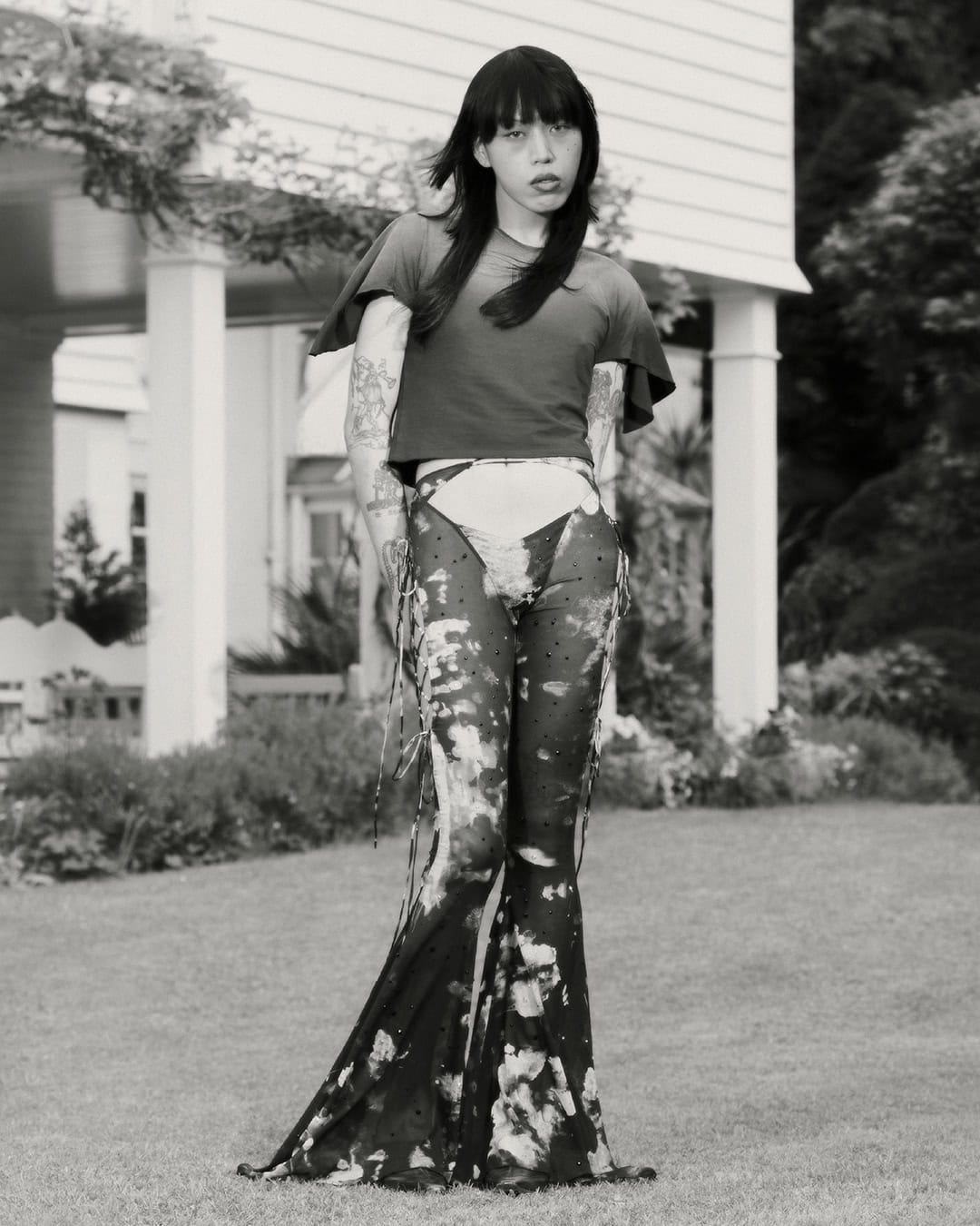 A person with long dark hair stands in a yard wearing avant-garde fashion, featuring a t-shirt and transparent, patterned pants. 