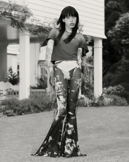 A person with long dark hair stands in a yard wearing avant-garde fashion, featuring a t-shirt and transparent, patterned pants. 