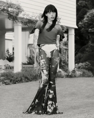 A person with long dark hair stands in a yard wearing avant-garde fashion, featuring a t-shirt and transparent, patterned pants. 