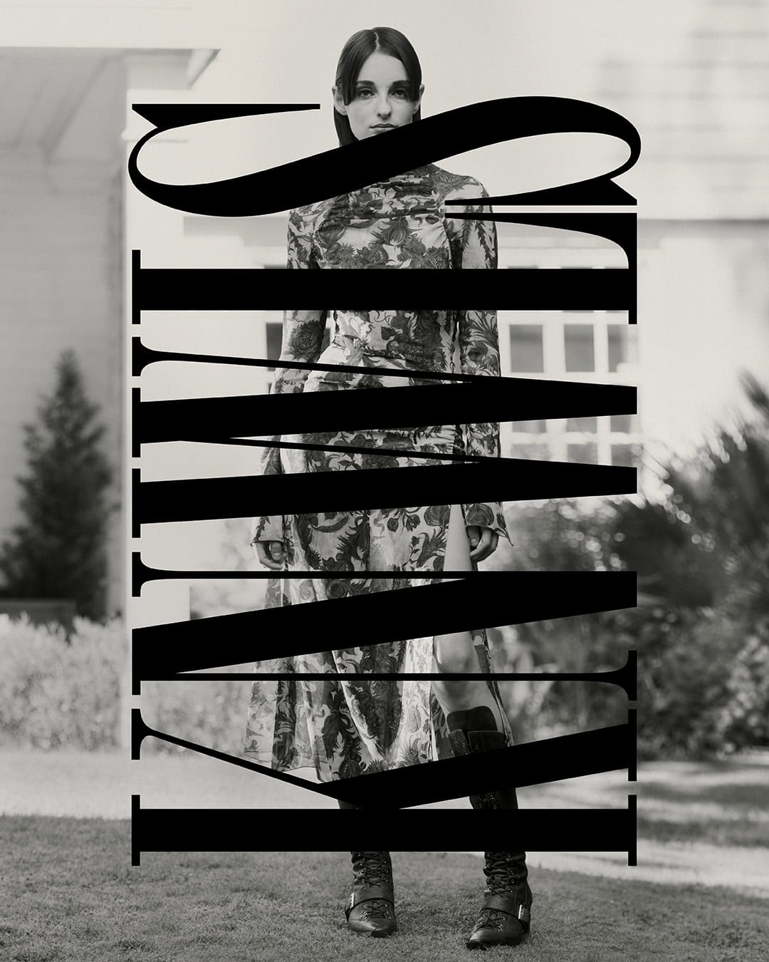 Black and white fashion shot of a woman in a floral dress and buckled boots standing outside a white house with an expansive lawn.