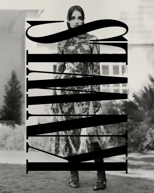 Black and white fashion shot of a woman in a floral dress and buckled boots standing outside a white house with an expansive lawn.