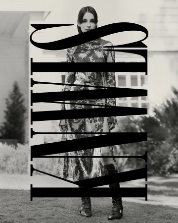 Black and white fashion shot of a woman in a floral dress and buckled boots standing outside a white house with an expansive lawn.