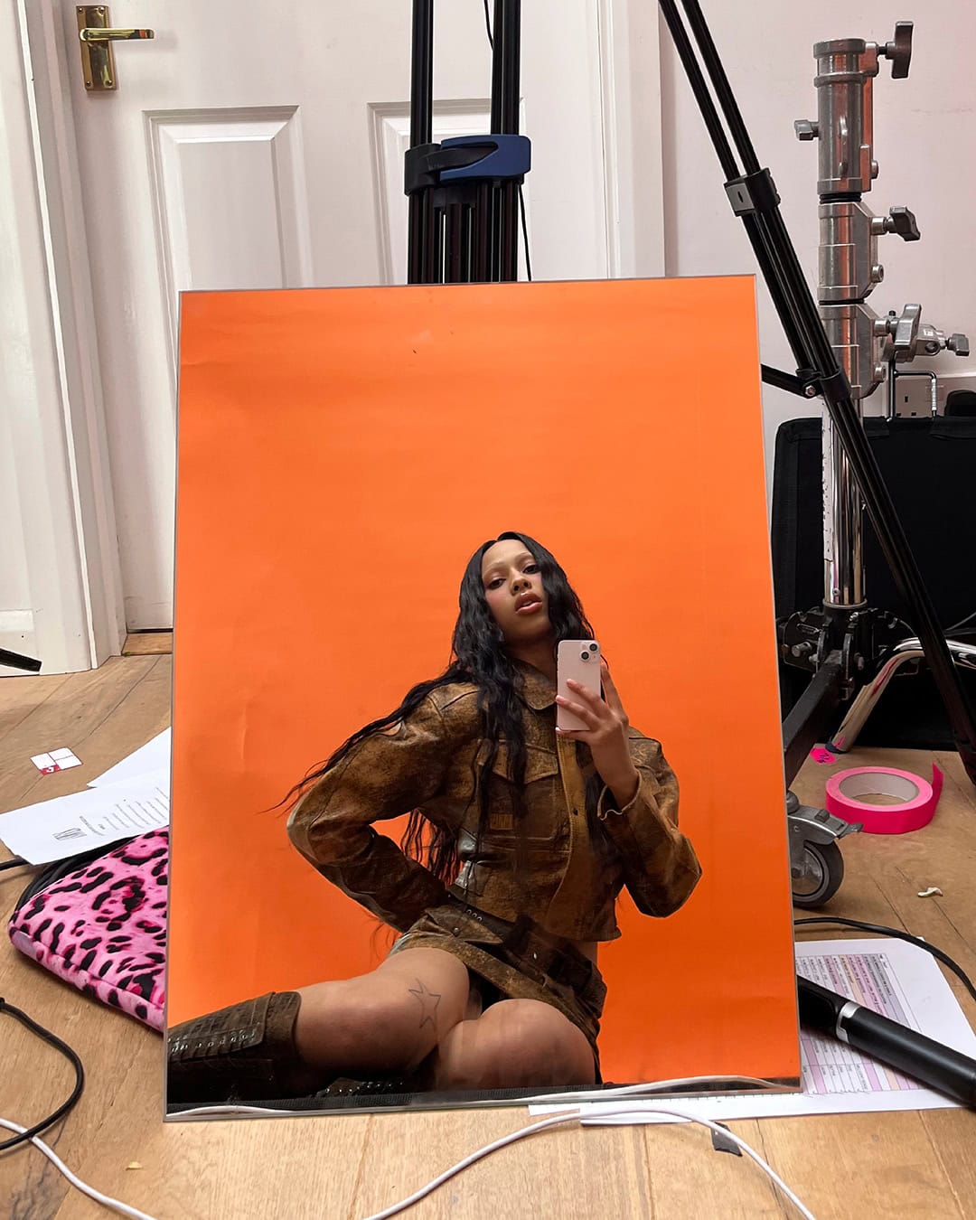 Woman in brown leather-like outfit takes selfie against orange backdrop in studio setting with film equipment visible.