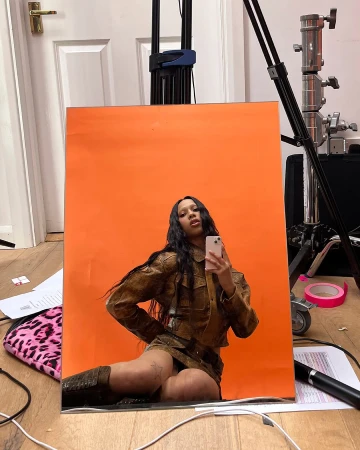 Woman in brown leather-like outfit takes selfie against orange backdrop in studio setting with film equipment visible.