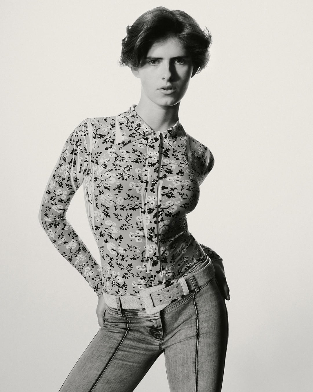 Monochrome portrait of a slender adult woman with short brown hair, posing in a floral blouse, jeans, and a wide belt against a light background.