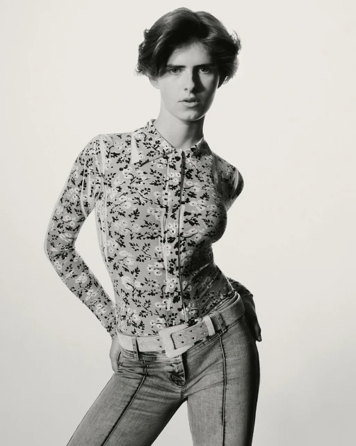 Monochrome portrait of a slender adult woman with short brown hair, posing in a floral blouse, jeans, and a wide belt against a light background.