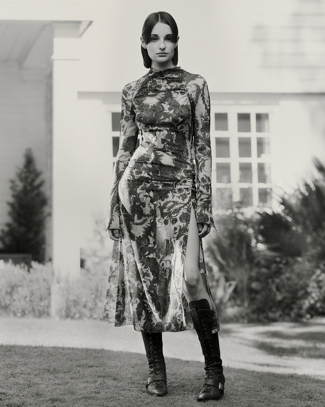 A slender woman in a floral midi dress and knee-high boots stands on grass with a neutral building in the background.