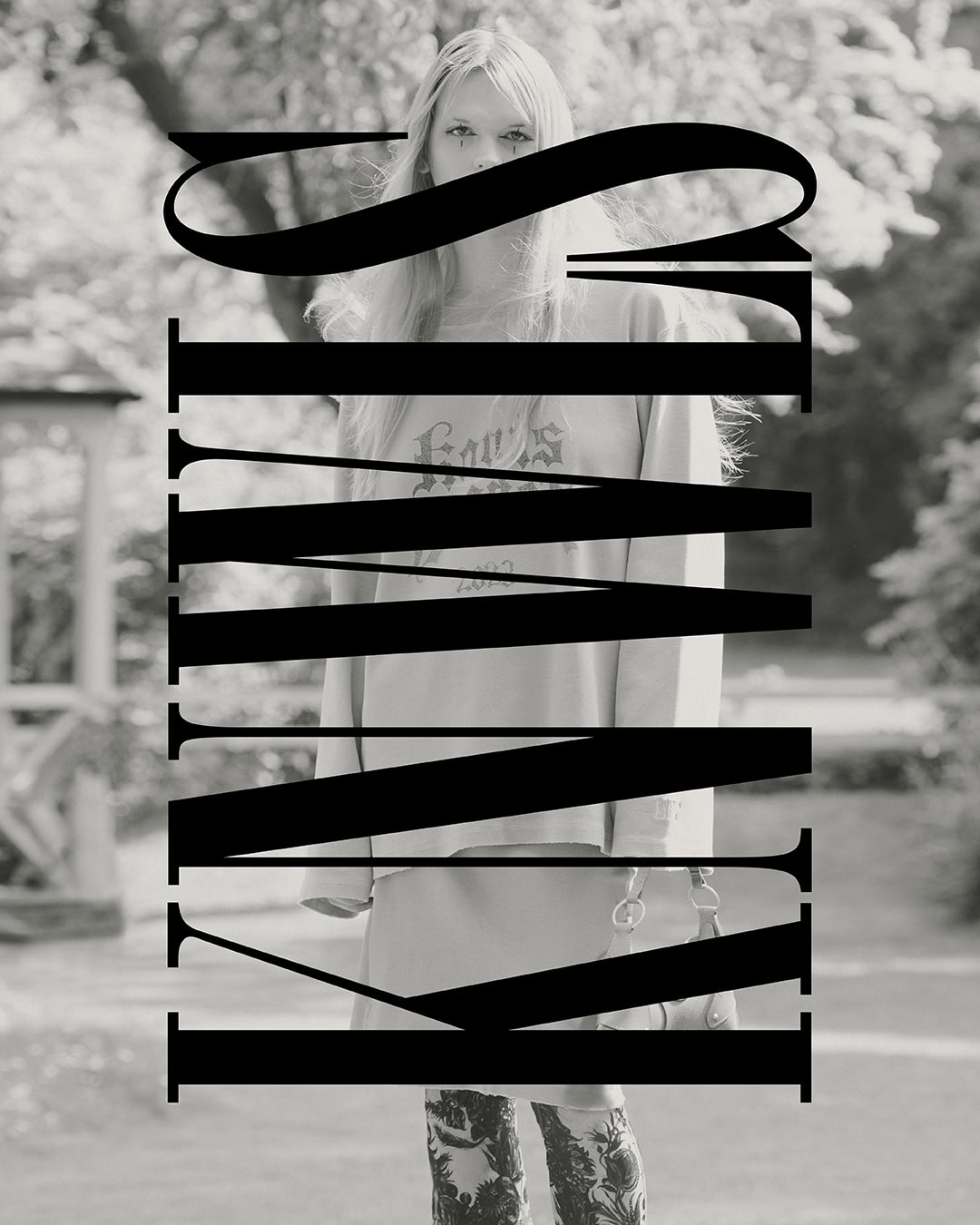 Black and white portrait of a woman with long blonde hair, a sweatshirt and patterned pants, standing outside with trees in the background.
