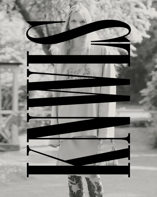 Black and white portrait of a woman with long blonde hair, a sweatshirt and patterned pants, standing outside with trees in the background.