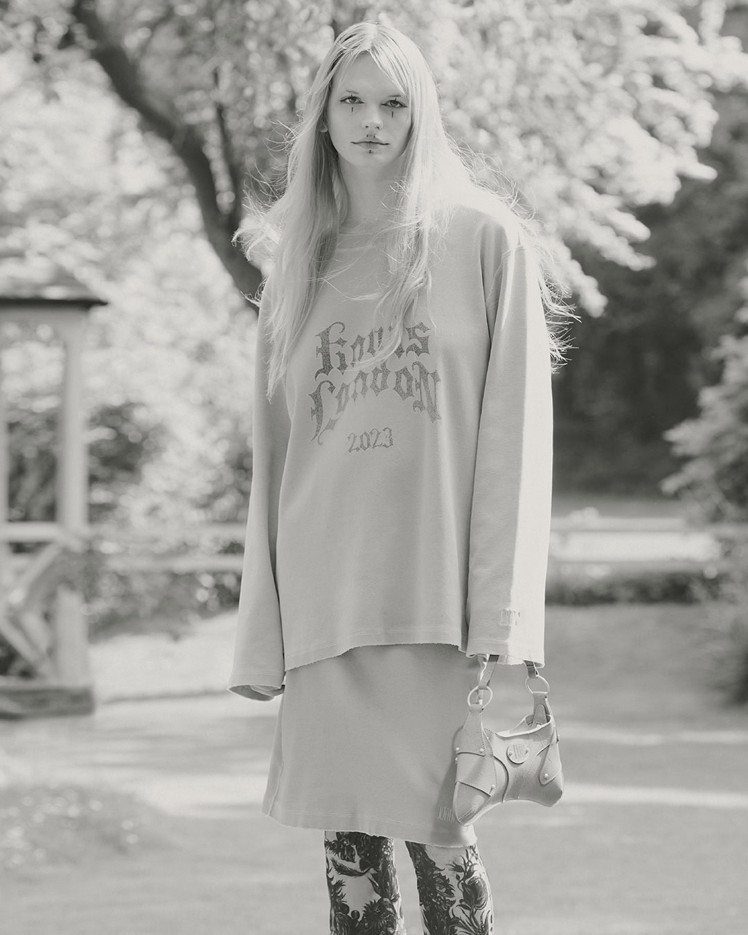 Monochrome outdoor fashion shot: young woman in long-sleeve shirt, skirt, patterned tights, holding purse in neutral light.