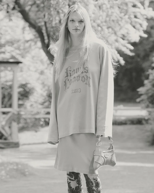 Monochrome outdoor fashion shot: young woman in long-sleeve shirt, skirt, patterned tights, holding purse in neutral light.