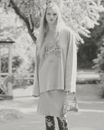 Monochrome outdoor fashion shot: young woman in long-sleeve shirt, skirt, patterned tights, holding purse in neutral light.