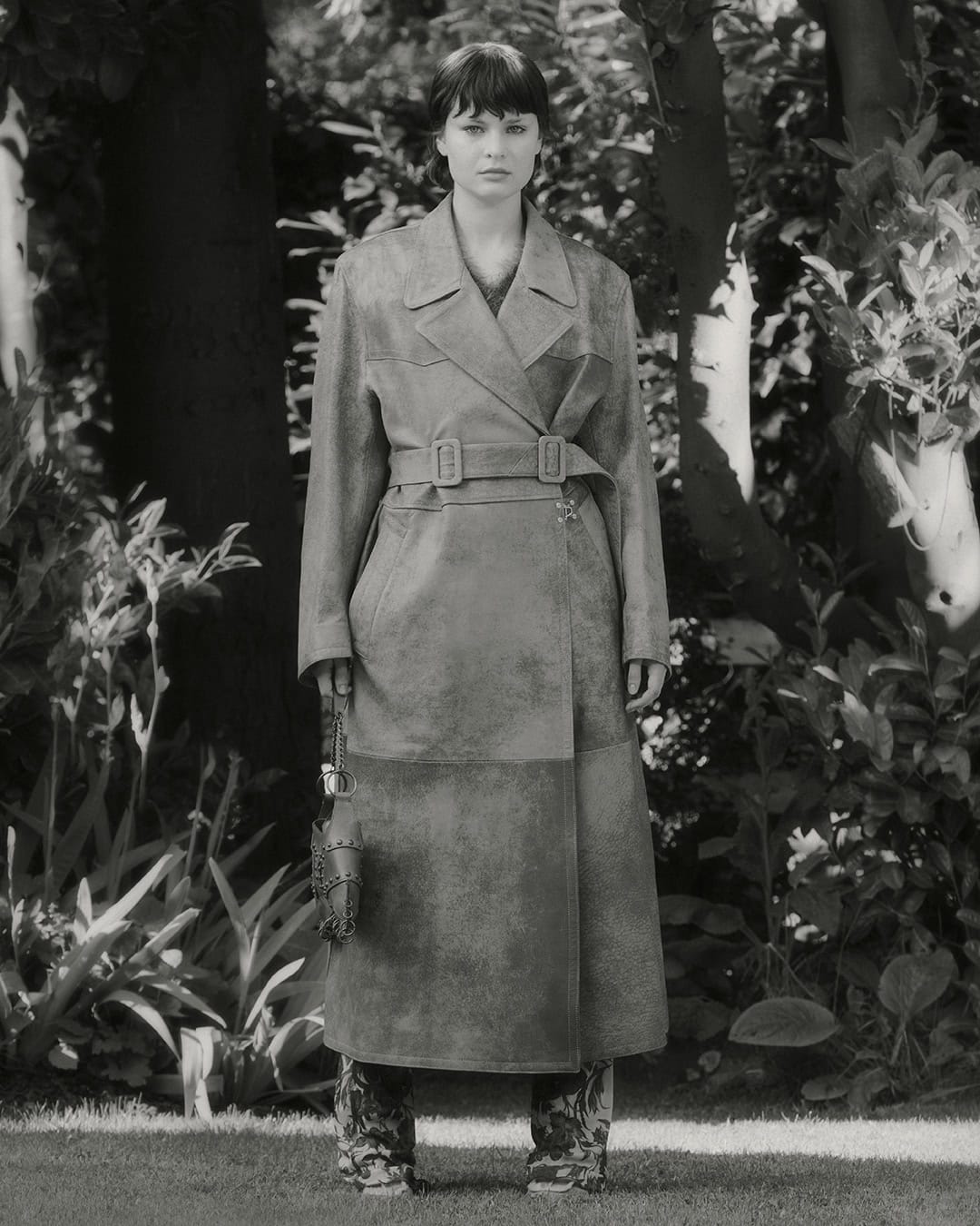 A young adult woman stands in an outdoor setting, modeling a long gray trench coat, with trees and greenery visible in the background.