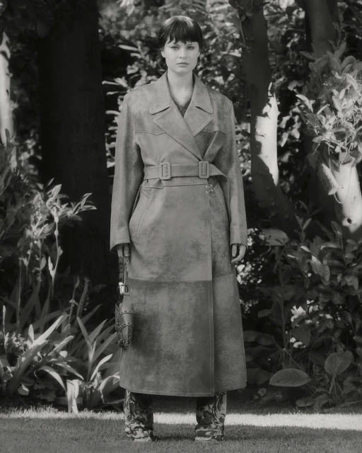 A young adult woman stands in an outdoor setting, modeling a long gray trench coat, with trees and greenery visible in the background.