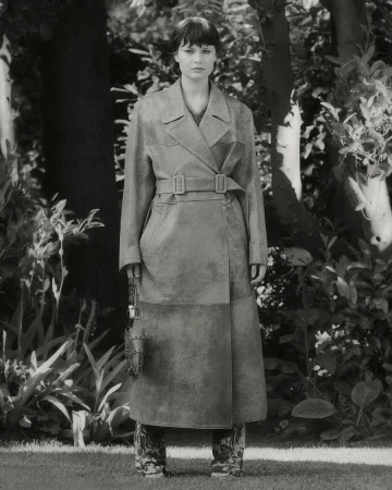 A young adult woman stands in an outdoor setting, modeling a long gray trench coat, with trees and greenery visible in the background.